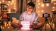 Image is a warm, cozy indoor scene featuring a young person with light skin and short, dark hair, sitting cross-legged on the floor. They are wearing a light gray sweatshirt and are smiling with delight as they open a glowing box of I Miss You Gifts in front of them. The room is softly illuminated by string lights draped around, creating a festive atmosphere. In the background, there are plush toys, including a teddy bear with a pink scarf, and framed pictures on the walls. A smartphone on a small tripod is positioned nearby, possibly recording the moment. The overall ambiance is cheerful and intimate, with a focus on the subject's joyful expression and the warm lighting that enhances the cozy setting.