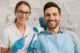 Smiling patient in dentist chair receiving tooth whitening treatment. Dentist applies gel and uses LED lamp in clean clinic.